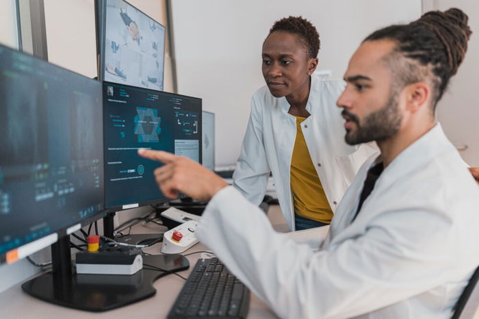 Two scientists in lab coats discuss data displayed on computer screens in a laboratory setting.